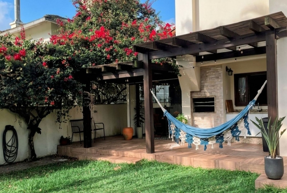 Varanda posterior com churrasqueira integrada ao amplo gramado, piscina e solarium em casa no Parque São Jorge, Itacorubi.