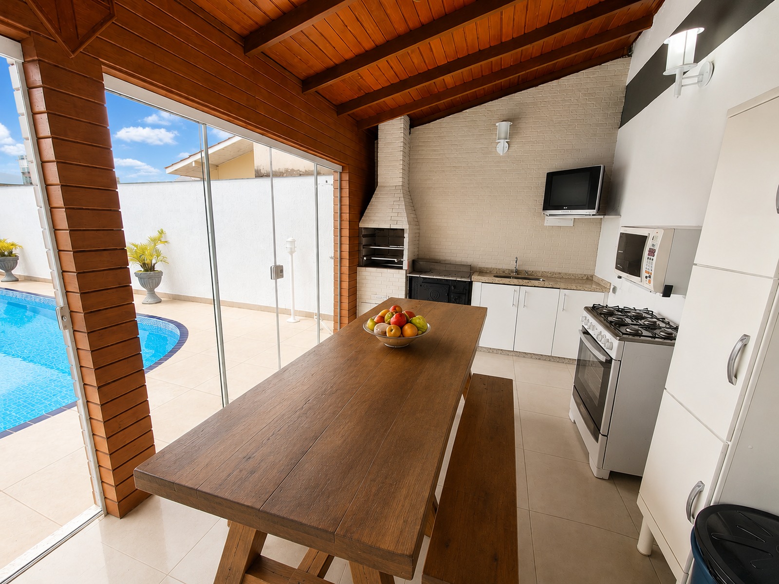 Espaço gourmet coberto com churrasqueira, bancada de apoio, mesa ampla e fechamento em vidro junto à piscina.