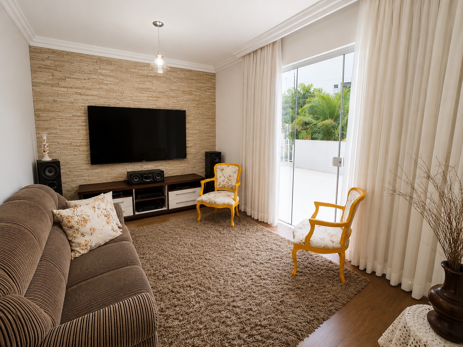 Sala íntima com painel de pedra natural, TV e ampla porta de vidro com acesso à área externa da residência (terraço). Vista para a cidade.